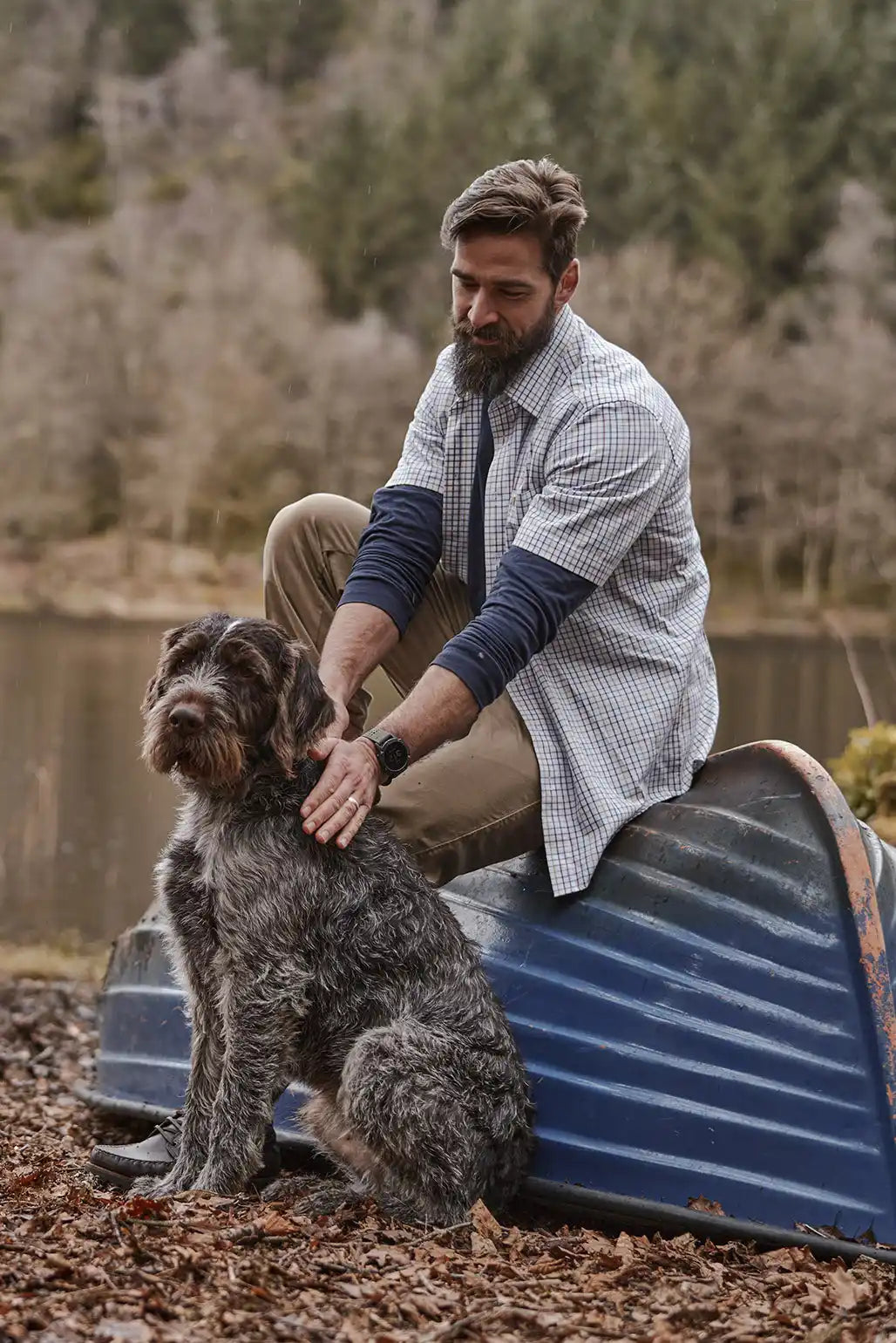 A man with a beard petting a Wirehaired Pointing Griffon dog while sitting on an overturned blue rowboat.