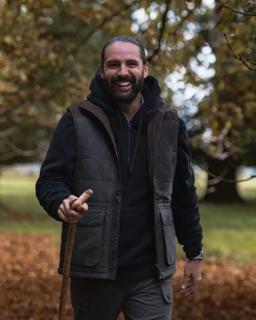 Smiling man in forest polar gilet with quilted vest, hoodie, cargo pants, holding a walking stick