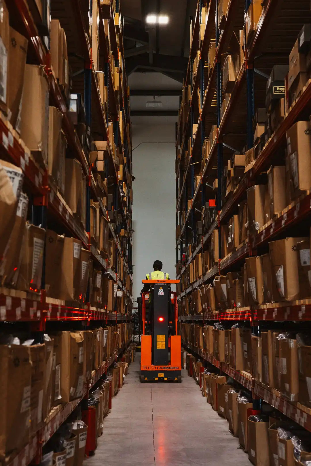 An orange and black reach truck with a person operating it, surrounded by rows of shelved boxes.