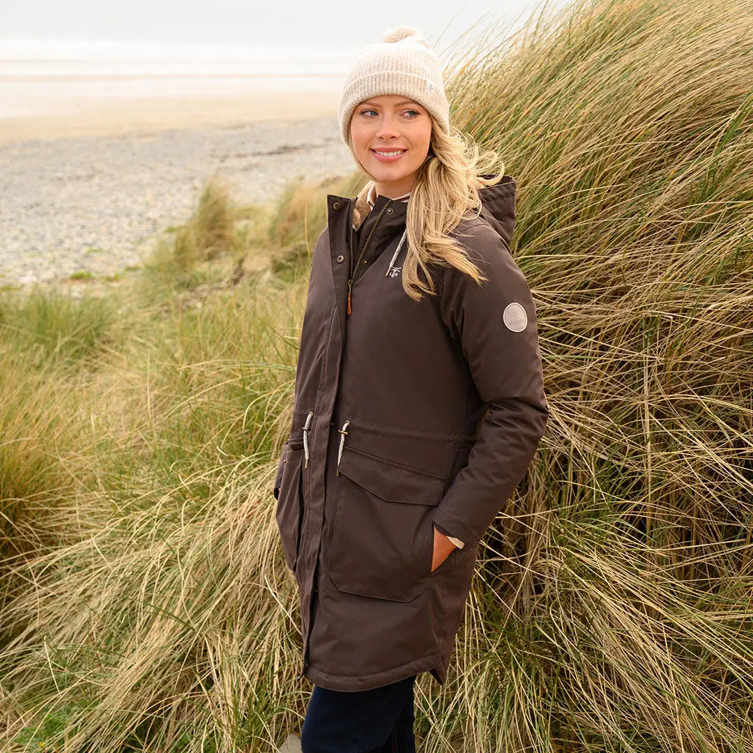 Woman in brown Lighthouse Isobel Coat and cream beanie outdoors near tall grass