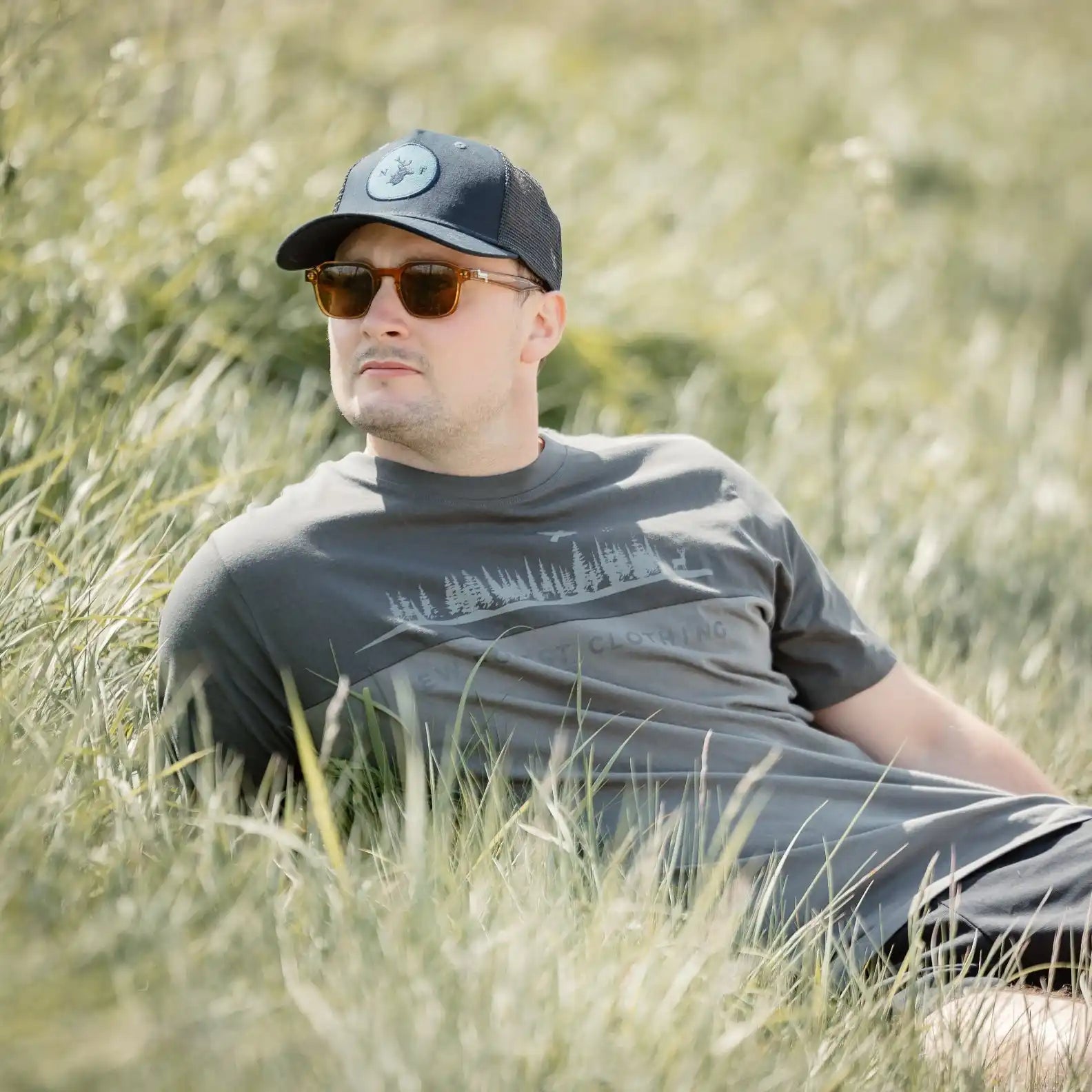 A man in a dark grey t-shirt, brown sunglasses, and a black baseball cap, relaxing in tall grass.