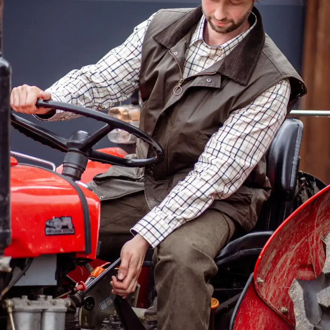 Man operating a red tractor wearing forest bushman wax gilet in outdoor farm scene