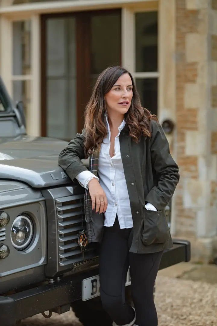 Woman leaning on gray SUV wearing forest freya ladies wax jacket and black pants