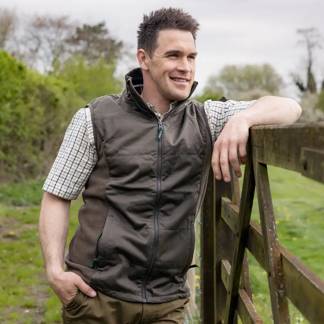 Man in dark vest over checkered shirt casually leans on fence showing Forest Grouse Padded Gilet