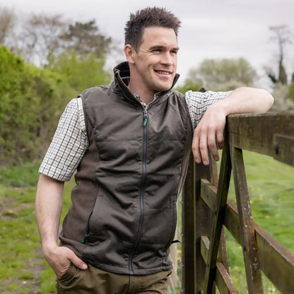Man in dark vest over checkered shirt casually leans on fence showing Forest Grouse Padded Gilet