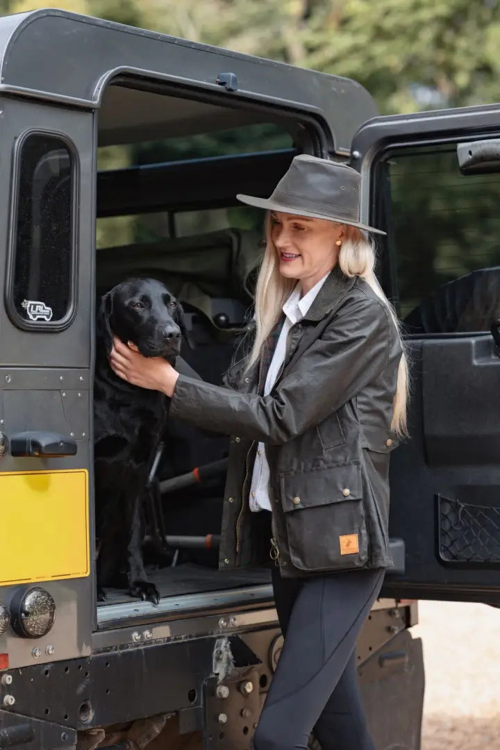 Blonde woman in a British Millerain wax jacket petting a black Labrador in forest clothing style