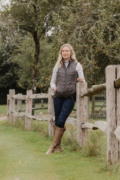 Woman in vest and boots leaning on a fence, wearing the New Forest Ladies Heated Gilet