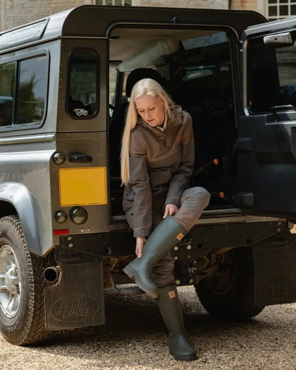 Woman in outdoor gear putting on green wellington boots wearing the Forest Nightjar Smock