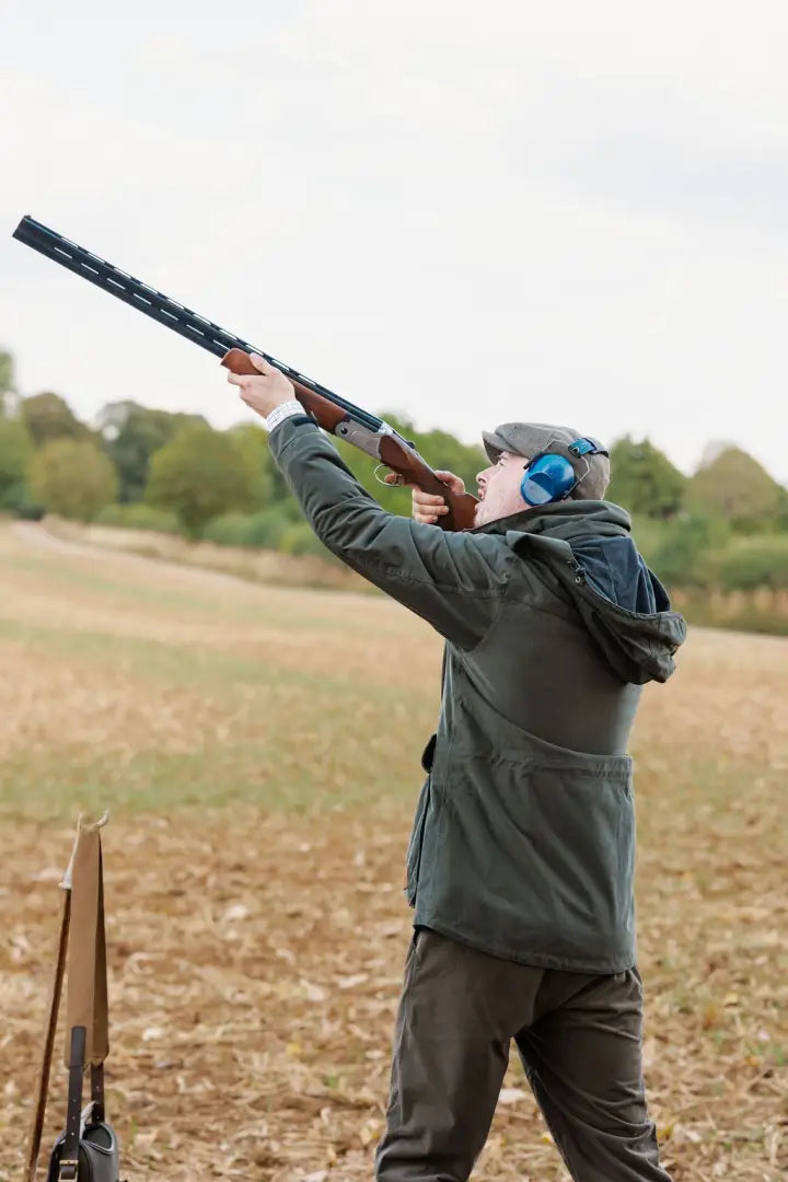Person aiming shotgun in forest marsh jacket with ear protection and cap