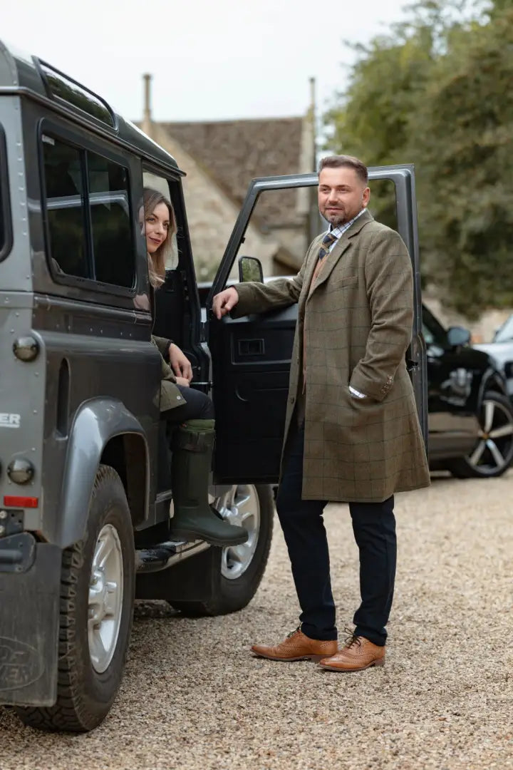 Man in forest moleskin jeans leaning on gray SUV door with woman inside outdoor style