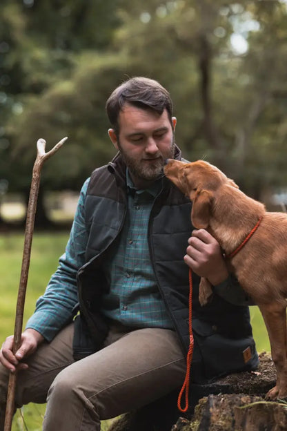 Man with beard wearing plaid shirt and forest polar gilet with brown dog licking face