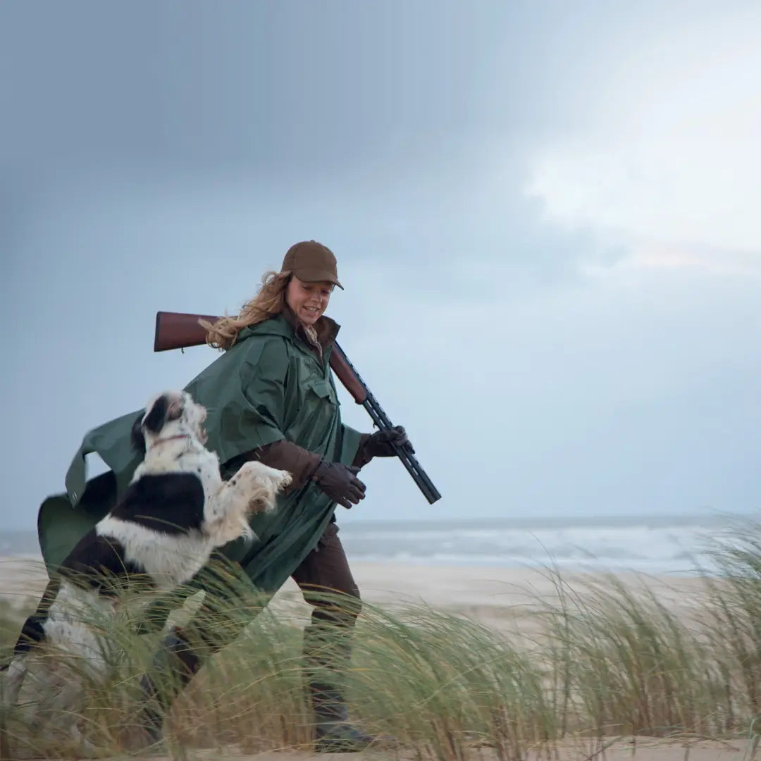 Hunter with a shotgun and dog on coastal grassland wearing a Percussion Waterproof Poncho
