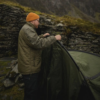 Person setting up a green tent in a rugged outdoor spot with a Snugpak Sleeka Elite Jacket