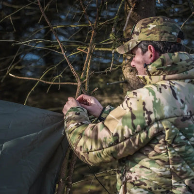 Soldier in camouflage using branches with Snugpak Stasha for wild camping weather shelter