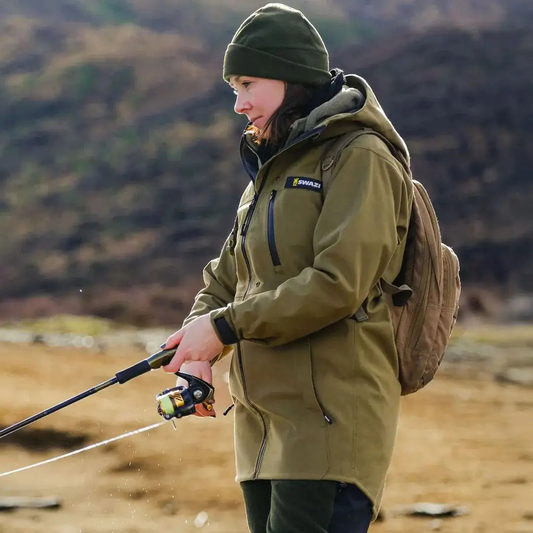 Woman in olive green Swazi Artemis Womens Jacket with hat and backpack holding a fishing rod, perfect for hunting and country clothing