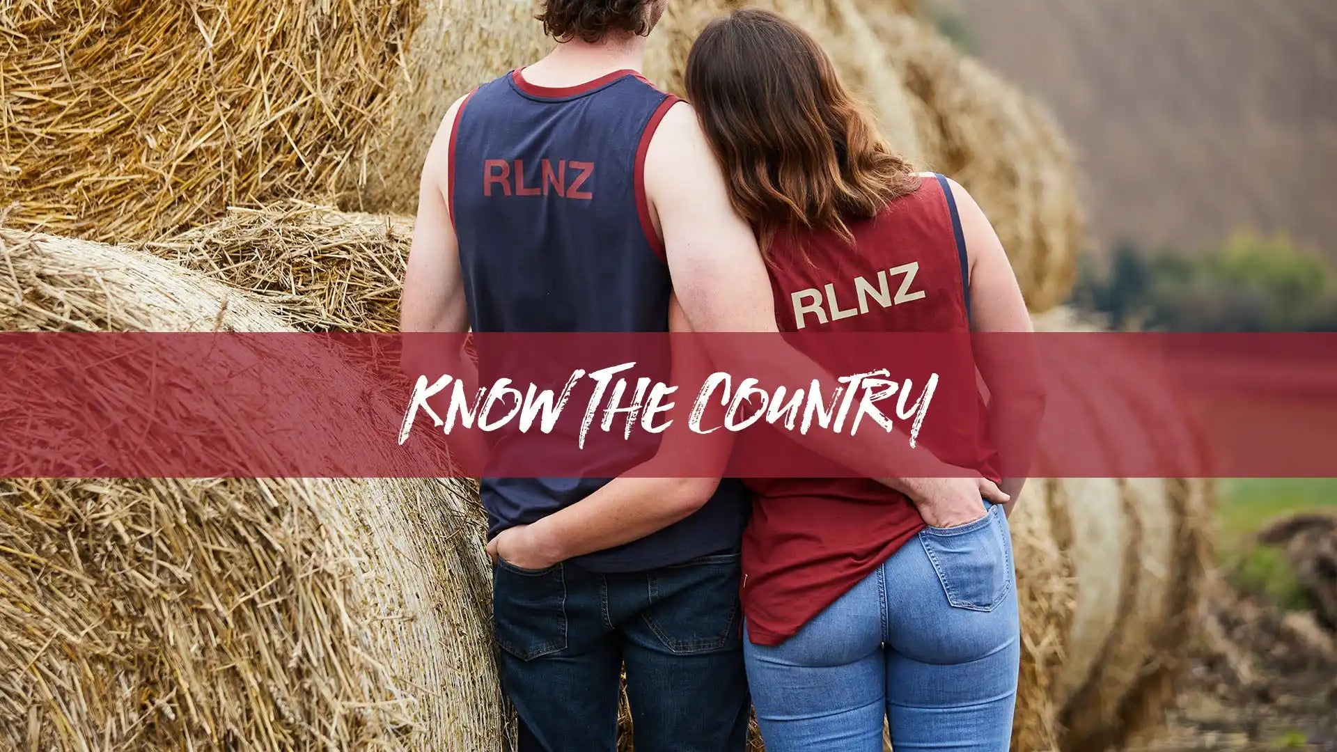 Two people wearing tank tops and jeans, embracing in front of hay bales.